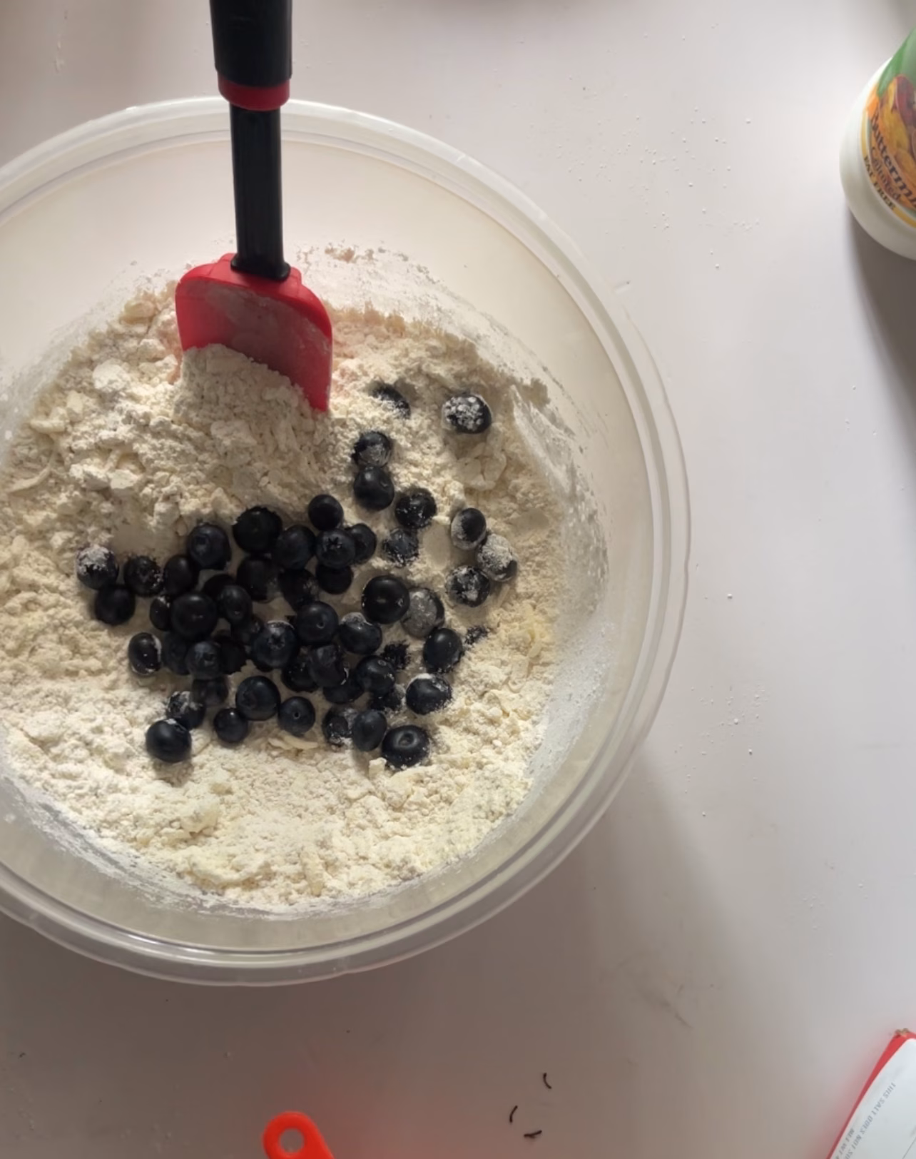 overhead shot of a clear plastic bowl of flour and butter mix with visible blueberries and black and red rubber spatula